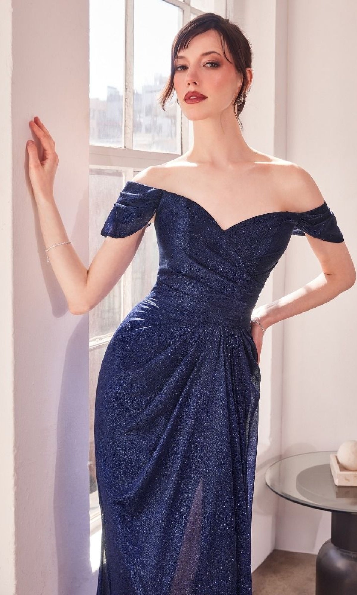 A woman in the Off-the-Shoulder Glitter Prom Dress: Amoris T604 stands by a sunlit window, one hand on the wall and the other on her hip.
